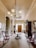 An elegant, vintage hallway features ornate wooden furniture, including armchairs with pink upholstery and a central round table. The room is adorned with a grand chandelier hanging from the ceiling. The walls are decorated with framed pictures and a large mirror. There is rich woodwork, with cabinets and a tall grandfather clock situated near the closed double doors at the end of the hallway. A long decorative rug runs along the wooden floor.