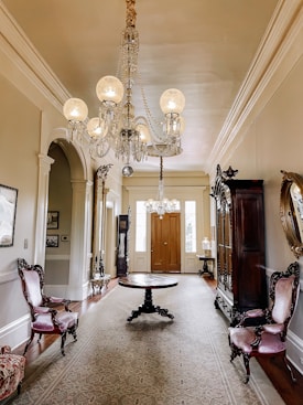 An elegant, vintage hallway features ornate wooden furniture, including armchairs with pink upholstery and a central round table. The room is adorned with a grand chandelier hanging from the ceiling. The walls are decorated with framed pictures and a large mirror. There is rich woodwork, with cabinets and a tall grandfather clock situated near the closed double doors at the end of the hallway. A long decorative rug runs along the wooden floor.