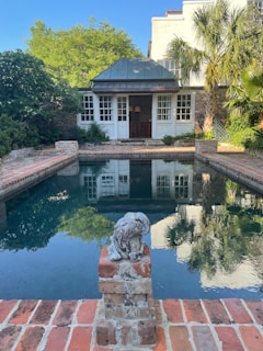 A small backyard pool surrounded by brick paving sits in front of a charming building with multiple windows and a dark roof. Trees and plants add lush greenery around the pool area. A decorative stone sculpture is positioned at the edge of the pool.
