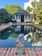 A small backyard pool surrounded by brick paving sits in front of a charming building with multiple windows and a dark roof. Trees and plants add lush greenery around the pool area. A decorative stone sculpture is positioned at the edge of the pool.