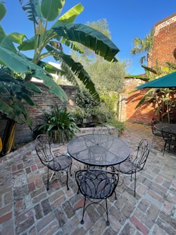 A patio area featuring a round metal table and several matching chairs on a brick surface. Lush tropical plants, including large banana leaves, surround the space. A brick wall and a clear blue sky are visible in the background, and part of a green umbrella can be seen to the right.