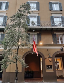 A classic building facade with multiple windows featuring blue shutters and decorative balconies. An American flag hangs prominently next to an arched entrance. A tree is positioned in the foreground, and a sign for 'Hotel Mazarin' is displayed beside a traditional lantern.