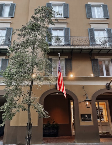 A classic building facade with multiple windows featuring blue shutters and decorative balconies. An American flag hangs prominently next to an arched entrance. A tree is positioned in the foreground, and a sign for 'Hotel Mazarin' is displayed beside a traditional lantern.