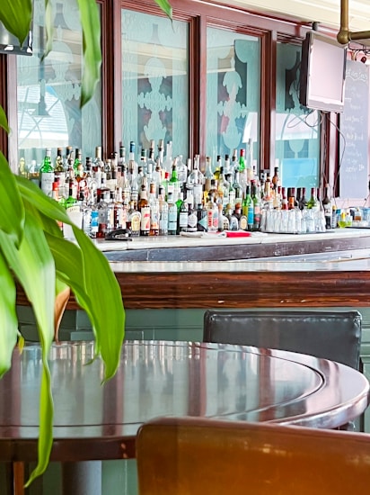 A bar counter with an extensive array of liquor bottles lined up against a stylish wooden panel with frosted glass designs. In front of the counter, a table with chairs is partially visible, along with some green plant leaves hanging down.