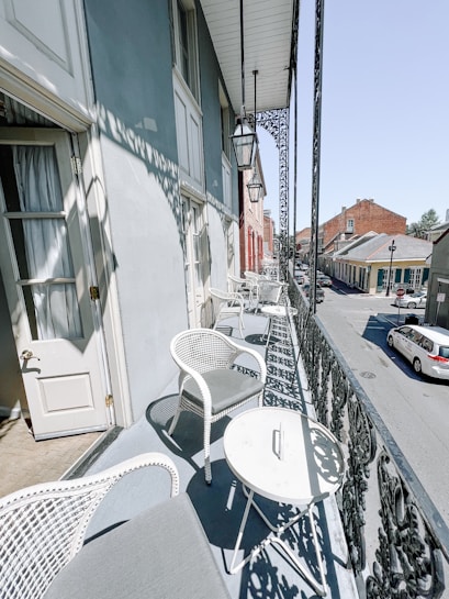 A narrow balcony lined with white wicker chairs and round tables overlooks a quiet urban street. Elegant wrought iron railings add a touch of classic charm, while sunlight creates intricate shadow patterns. A street with parked cars and historic-style buildings can be seen in the background.
