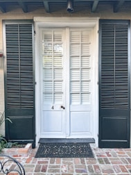 A brick pathway leads to a white door with a traditional design, flanked by two dark green shutters. The door features glass panels and horizontal wooden slats. A black ornamental doormat lies in front of the entrance, contrasting with the red and brown bricks. A few green plants are visible on the left side.