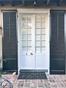 A brick pathway leads to a white door with a traditional design, flanked by two dark green shutters. The door features glass panels and horizontal wooden slats. A black ornamental doormat lies in front of the entrance, contrasting with the red and brown bricks. A few green plants are visible on the left side.
