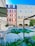 A serene courtyard of a hotel with a central swimming pool surrounded by lounge chairs. The architecture includes a combination of modern and classical design elements, with brick and stucco walls and red shutters. Lush green plants add a touch of nature to the urban setting.