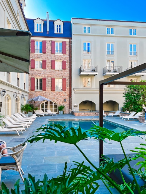 A serene courtyard of a hotel with a central swimming pool surrounded by lounge chairs. The architecture includes a combination of modern and classical design elements, with brick and stucco walls and red shutters. Lush green plants add a touch of nature to the urban setting.