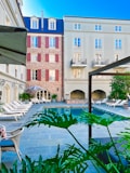 A serene courtyard of a hotel with a central swimming pool surrounded by lounge chairs. The architecture includes a combination of modern and classical design elements, with brick and stucco walls and red shutters. Lush green plants add a touch of nature to the urban setting.