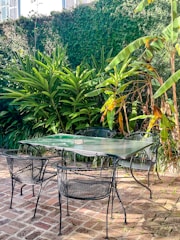 A glass-top table with four metal mesh chairs is placed on a rustic brick patio. The area is surrounded by lush green foliage, including tall tropical plants and a leafy wall in the background. Sunlight creates a play of light and shadow on the ground and furniture.