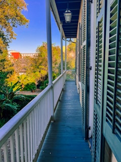 A wooden balcony with blue painted flooring and white railings extends along the side of a building. Green shutters are open along the exterior wall, and overhead, a series of lantern-style lights hang. Lush, green foliage and various trees surround the area, with a distant red brick building visible against a vibrant blue sky.