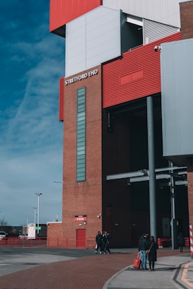 A large building structure with red and white elements prominently features the label 'Stretford End'. Several people are walking near the base of the building, and one person is pulling a red suitcase. The sky is mostly clear with a few clouds, adding a bright atmosphere to the overall urban scene.