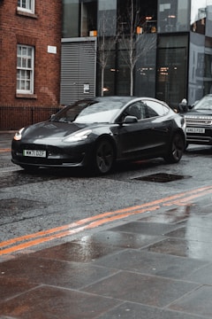 A sleek, black Tesla car is parked on a wet street in an urban area. Behind the Tesla, another car is partially visible. The street appears to be empty of pedestrians and is flanked by a brick building on one side and a modern glass building on the other. The pavement is wet, reflecting the overcast weather.