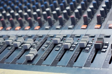 A close-up view of an audio mixing console with various sliders and knobs arranged in rows. The equipment appears complex, used for adjusting sound levels and mixing audio. The sliders are positioned at different levels, suggesting active sound management.