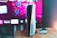 A gaming console is placed on a wooden table with a game controller nearby. In the background, a television screen displays colorful graphics, including geometric shapes and vibrant colors.