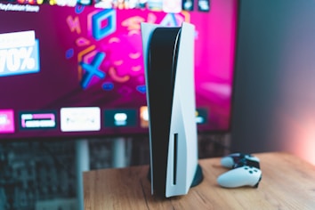 A gaming console is placed on a wooden table with a game controller nearby. In the background, a television screen displays colorful graphics, including geometric shapes and vibrant colors.