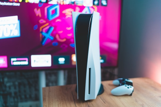 A gaming console is placed on a wooden table with a game controller nearby. In the background, a television screen displays colorful graphics, including geometric shapes and vibrant colors.