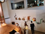 Inside a brewery, a large copper brewing vat is visible in the foreground. On the tiled wall behind it, there is a detailed flowchart depicting different stages of the brewing process, including malting, lautering, and fermenting. Several people are observing the installation, with some standing close to the machinery and others on an elevated platform in the background.