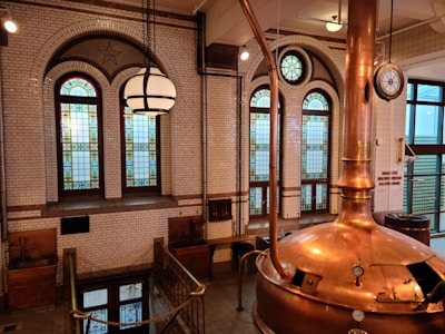 An interior scene of a traditional brewery with stained glass windows set in a brick wall. A large copper brewing kettle with pipes and gauges is prominently featured in the foreground. The warm lighting and intricate details of the stained glass create a historic and industrial atmosphere.