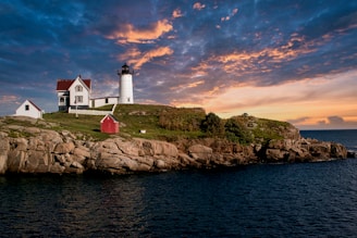A picturesque lighthouse sits atop a rocky shoreline with two adjacent buildings, surrounded by green grass. The sky is filled with dramatic clouds illuminated by the warm hues of a setting sun.