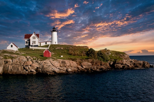 A picturesque lighthouse sits atop a rocky shoreline with two adjacent buildings, surrounded by green grass. The sky is filled with dramatic clouds illuminated by the warm hues of a setting sun.