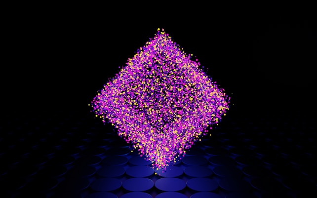 A geometric shape composed of numerous small, colorful particles floats in a dark environment. The particles are primarily pink, purple, and yellow, forming a diamond-like structure. The background features a pattern of dark circular shapes that extend into the distance.