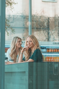 Two women with long hair are sitting at a table in a cafe. One is holding a coffee cup, and both appear to be smiling and engaged in conversation. There are shelves in the background stocked with various brightly colored beverages. The cafe is well-lit with a modern interior design.