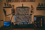 A home office setup featuring two computer monitors with abstract line art wallpaper, flanked by two wall-mounted speakers. A gaming PC with illuminated components is on the right, and a laptop displaying a nature scene is on the left. The desk is equipped with a microphone arm and various tech gadgets, including a keyboard and a small tablet. Shelves above hold action figures and framed posters.