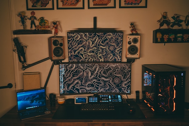 A home office setup featuring two computer monitors with abstract line art wallpaper, flanked by two wall-mounted speakers. A gaming PC with illuminated components is on the right, and a laptop displaying a nature scene is on the left. The desk is equipped with a microphone arm and various tech gadgets, including a keyboard and a small tablet. Shelves above hold action figures and framed posters.