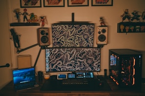 A home office setup featuring two computer monitors with abstract line art wallpaper, flanked by two wall-mounted speakers. A gaming PC with illuminated components is on the right, and a laptop displaying a nature scene is on the left. The desk is equipped with a microphone arm and various tech gadgets, including a keyboard and a small tablet. Shelves above hold action figures and framed posters.