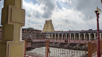 An ancient temple complex with intricate architecture that includes a prominent gopuram (tower) in the background. The compound includes a large open area with a water body surrounded by a fence. Cloudy skies can be seen above, casting a serene atmosphere over the scene.