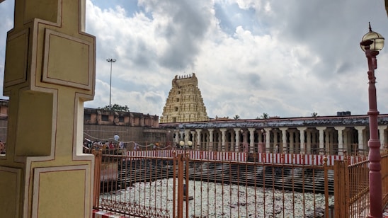 An ancient temple complex with intricate architecture that includes a prominent gopuram (tower) in the background. The compound includes a large open area with a water body surrounded by a fence. Cloudy skies can be seen above, casting a serene atmosphere over the scene.