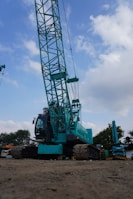 A large teal-colored construction crane is situated on a dirt surface under a partly cloudy sky. The crane features a prominent lattice boom and tracks for movement. Surrounding the crane are some trees and another smaller crane visible in the background.