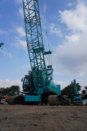 A large teal-colored construction crane is situated on a dirt surface under a partly cloudy sky. The crane features a prominent lattice boom and tracks for movement. Surrounding the crane are some trees and another smaller crane visible in the background.