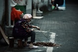 An elderly person wearing a patterned jacket and a headscarf sits on a small stool in an alleyway. They are poking at a small pile of sticks and debris with a stick. The surroundings include a wall with some Chinese characters, plastic bags, and a textured stone pavement.