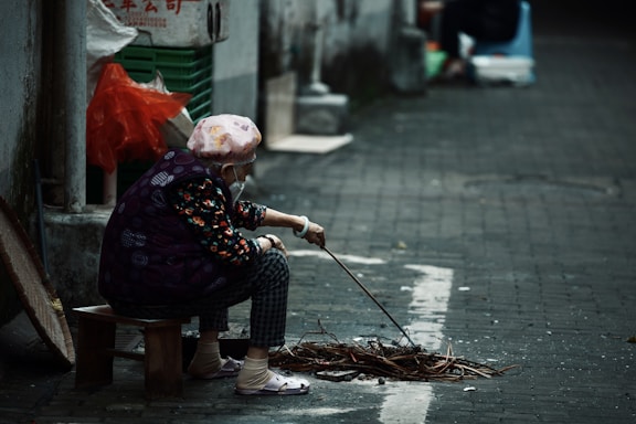An elderly person wearing a patterned jacket and a headscarf sits on a small stool in an alleyway. They are poking at a small pile of sticks and debris with a stick. The surroundings include a wall with some Chinese characters, plastic bags, and a textured stone pavement.