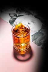 A beautifully crafted cocktail sits in a clear glass with large ice cubes and an elegant twist of lemon peel on top. The drink has a rich amber color, and the glass casts a shadow on a soft pink surface. In the background, there is a floral pattern featuring white flowers and green leaves, creating a sophisticated and artistic ambiance.