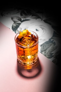 A beautifully crafted cocktail sits in a clear glass with large ice cubes and an elegant twist of lemon peel on top. The drink has a rich amber color, and the glass casts a shadow on a soft pink surface. In the background, there is a floral pattern featuring white flowers and green leaves, creating a sophisticated and artistic ambiance.