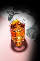 A beautifully crafted cocktail sits in a clear glass with large ice cubes and an elegant twist of lemon peel on top. The drink has a rich amber color, and the glass casts a shadow on a soft pink surface. In the background, there is a floral pattern featuring white flowers and green leaves, creating a sophisticated and artistic ambiance.