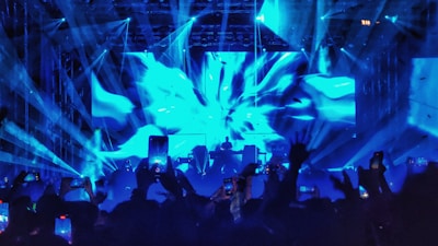 A vibrant concert scene with dynamic blue lights and laser beams illuminating the stage and audience. A DJ or performer is silhouetted against the bright backdrop, while the crowd is actively engaged, holding up phones to capture the moment.
