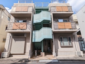 A three-story residential building with a modern design, featuring prominent balconies with diagonal wooden panels on each floor. A light blue staircase is centrally located, providing access to each level, and a bicycle is parked underneath the staircase. The building has a beige facade and is surrounded by other urban structures.