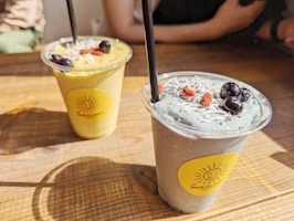 Two smoothie cups are placed on a wooden table, each with a black straw. The left smoothie is green and garnished with blueberries and possibly shredded coconut. The right smoothie has a light blueish-gray color, also garnished with blueberries, goji berries, and shredded coconut. Both cups have a bright yellow sticker with a sun logo.