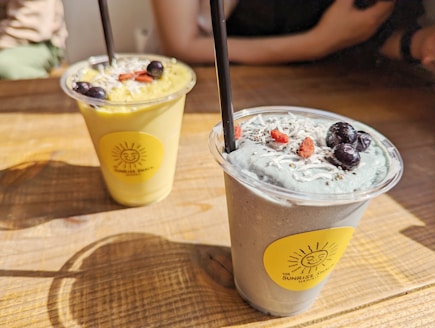 Two smoothie cups are placed on a wooden table, each with a black straw. The left smoothie is green and garnished with blueberries and possibly shredded coconut. The right smoothie has a light blueish-gray color, also garnished with blueberries, goji berries, and shredded coconut. Both cups have a bright yellow sticker with a sun logo.
