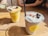 Two smoothie cups are placed on a wooden table, each with a black straw. The left smoothie is green and garnished with blueberries and possibly shredded coconut. The right smoothie has a light blueish-gray color, also garnished with blueberries, goji berries, and shredded coconut. Both cups have a bright yellow sticker with a sun logo.