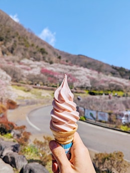 A hand holding a soft-serve ice cream cone with a swirl of light pink ice cream. The background features a scenic landscape with rolling hills covered in flowering trees in shades of pink and white. A curving road and some fencing are visible, under a clear blue sky.