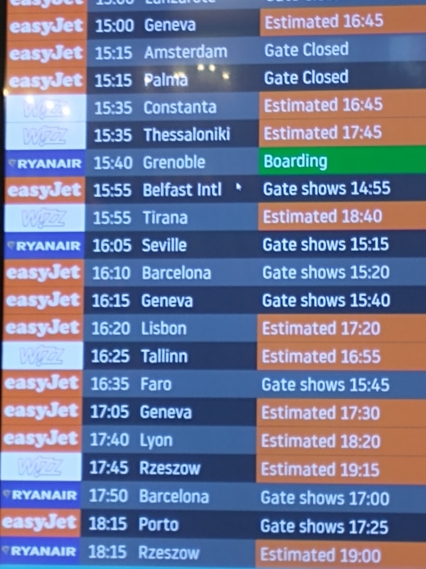 A flight departure board displaying various airlines and their scheduled flights. The board shows destinations, companies such as EasyJet, Ryanair, and Wizz, departure times, and status updates like 'Gate Closed', 'Estimated', or 'Boarding'. The background color for EasyJet is orange, Ryanair is blue, and Wizz is purple, with corresponding flight status updates in different colors.