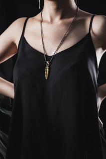 A person is wearing a black spaghetti strap top and a long necklace with a unique pendant shaped like a capsule. The person's arms are positioned akimbo, and the lighting emphasizes the sheen of the fabric.