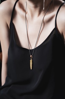 A person wearing a black spaghetti strap top is adorned with a necklace featuring a gold pendant in a unique design. The necklace has a sleek, modern aesthetic, adding elegance to the overall look.