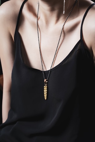 A person wearing a black spaghetti strap top is adorned with a necklace featuring a gold pendant in a unique design. The necklace has a sleek, modern aesthetic, adding elegance to the overall look.